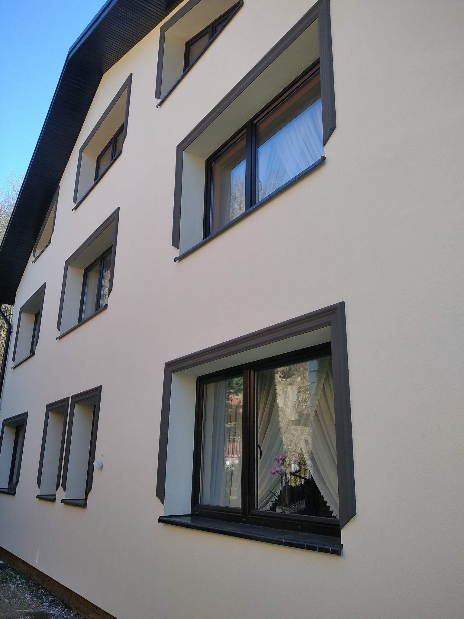 Elewacja budynku dwupiętrowego z oknami w ciemnych, profilowanych obramowaniach, widok z dołu na jasną fasadę w słoneczny dzień.