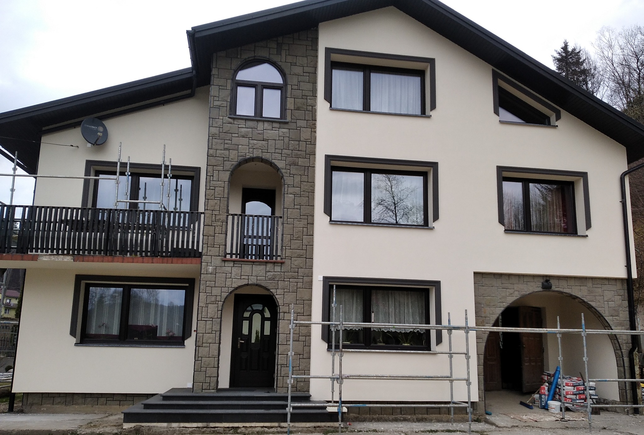 Dwupiętrowy dom z jasną elewacją i elementami dekoracyjnymi z kamienia, widoczne rusztowania wskazujące na prace wykończeniowe, na ścianie satelita.