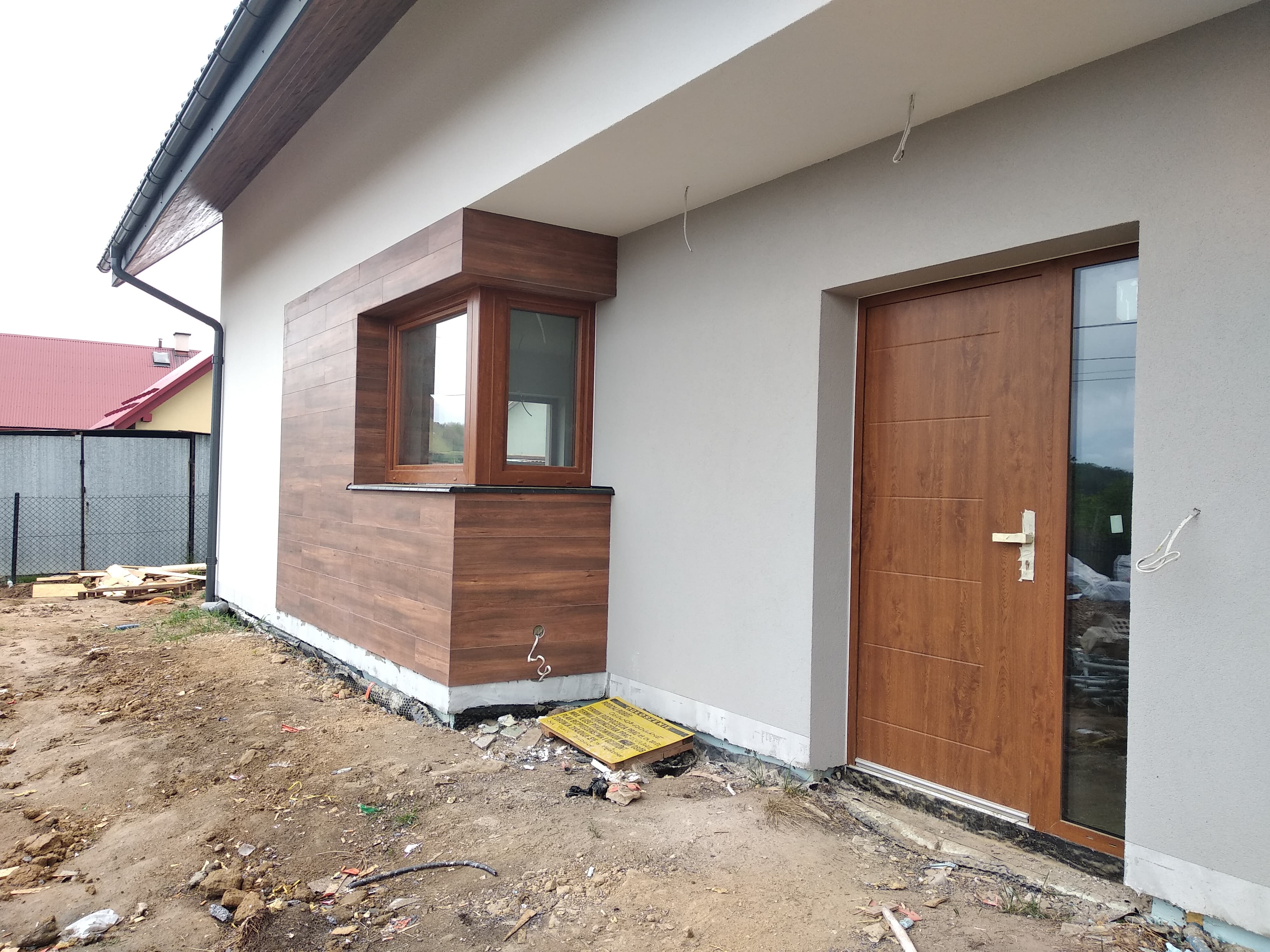 Fragment elewacji budynku z drewnopodobnymi płytami wokół okna i drewnianymi drzwiami wejściowymi; widoczne niedokończone prace ziemne wokół fundamentów.