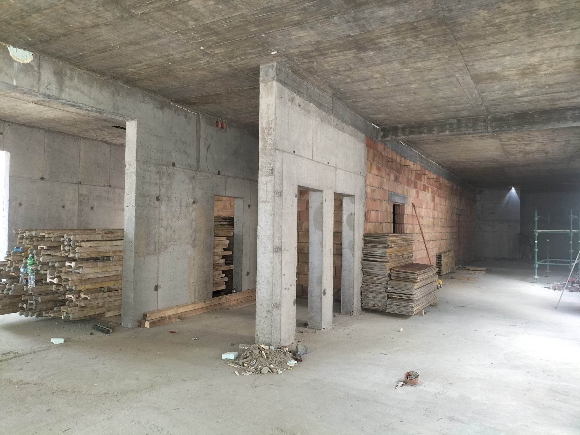 Wnętrze surowego budynku w trakcie budowy z widocznymi betonowymi ścianami, kolumnami i stropem, stertą drewnianych belek po lewej oraz stosem płyt po prawej stronie, z ceglaną ścianą w tle.