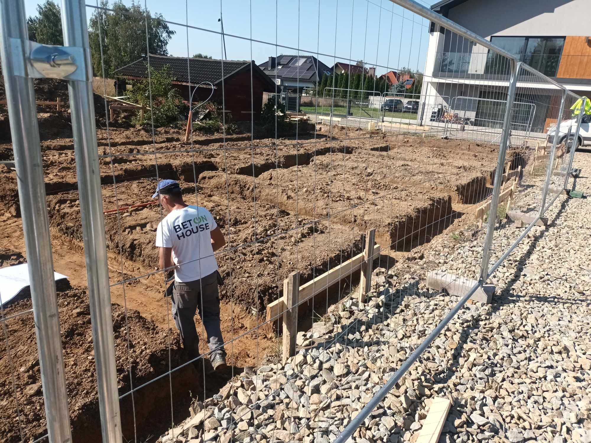 Wykopy pod fundamenty domu jednorodzinnego, widoczne szalunki drewniane i pracownik w koszulce z napisem 'BETON HOUSE', ogrodzenie tymczasowe.