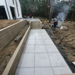 Ułożona ścieżka z jasnych płyt prowadząca do schodów, otoczona niskimi murkami oporowymi, w tle nowoczesny dom i las, widoczni pracownicy i ognisko z dymem.