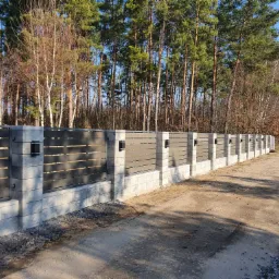 Długi, nowoczesny płot z szarych betonowych słupków i poziomych, ciemnoszarych paneli, oświetlony małymi lampami, biegnący wzdłuż polnej drogi i lasu w słoneczny dzień.