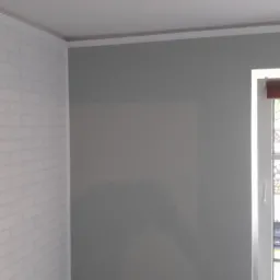 Pokój po remoncie: ściana pokryta tapetą imitującą białą cegłę, sąsiadująca z szarą ścianą przy oknie z brązową roletą.