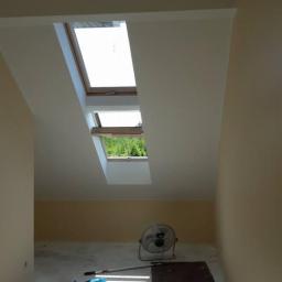 MalHus Szpachlowanie Natryskowe, Malowanie, GK Zabudowy,Poddasza - Pokój na poddaszu z dwoma oknami dachowymi, częściowo pomalowany na beżowo, z wentylatorem stojącym na podłodze i narzędziami malarskimi w rogu.