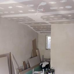 MalHus Szpachlowanie Natryskowe, Malowanie, GK Zabudowy,Poddasza - Pokój w trakcie montażu płyt kartonowo-gipsowych na suficie, widoczne łączenia płyt wypełnione masą szpachlową, na podłodze stoją płyty, drabina, worki z zaprawą i puszki z farbą.