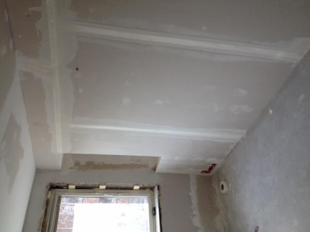 Ściana i sufit w trakcie prac wykończeniowych, widoczne ślady szpachlowania i gładzi, częściowo zamaskowane okno.