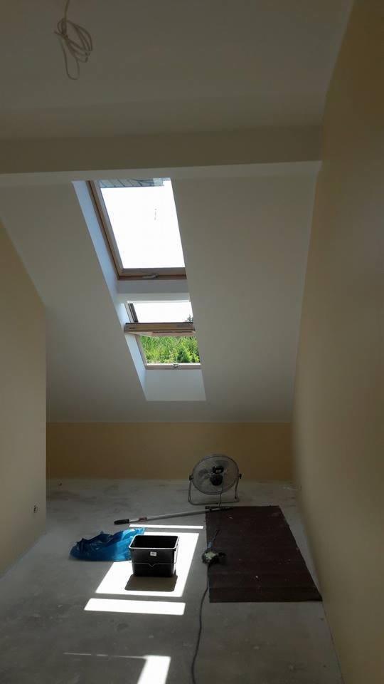 Pokój na poddaszu z dwoma oknami dachowymi, częściowo pomalowany na beżowo, z wentylatorem stojącym na podłodze i narzędziami malarskimi w rogu.