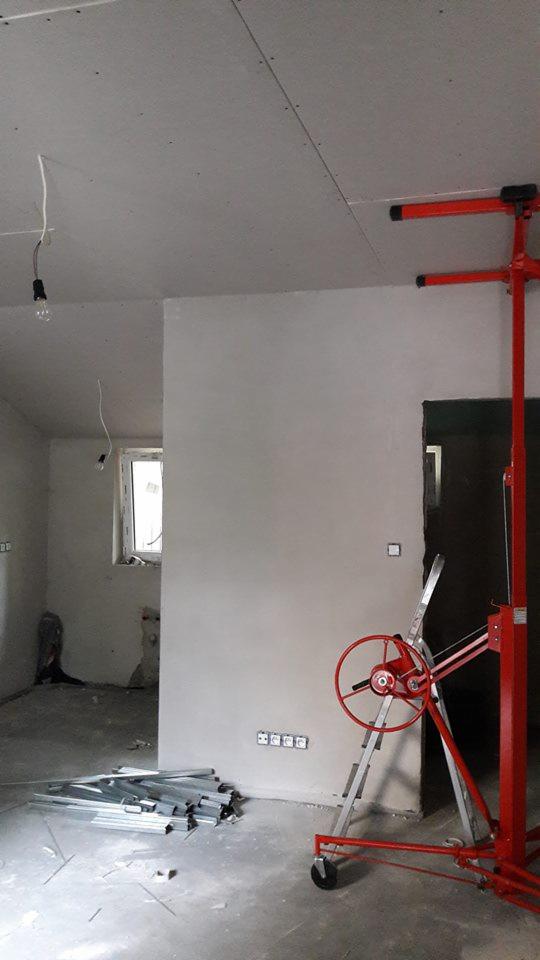 Wnętrze w trakcie prac wykończeniowych: zamontowane płyty kartonowo-gipsowe na suficie, ściana wstępnie wyrównana, widoczny podnośnik do płyt oraz profile konstrukcyjne na podłodze.