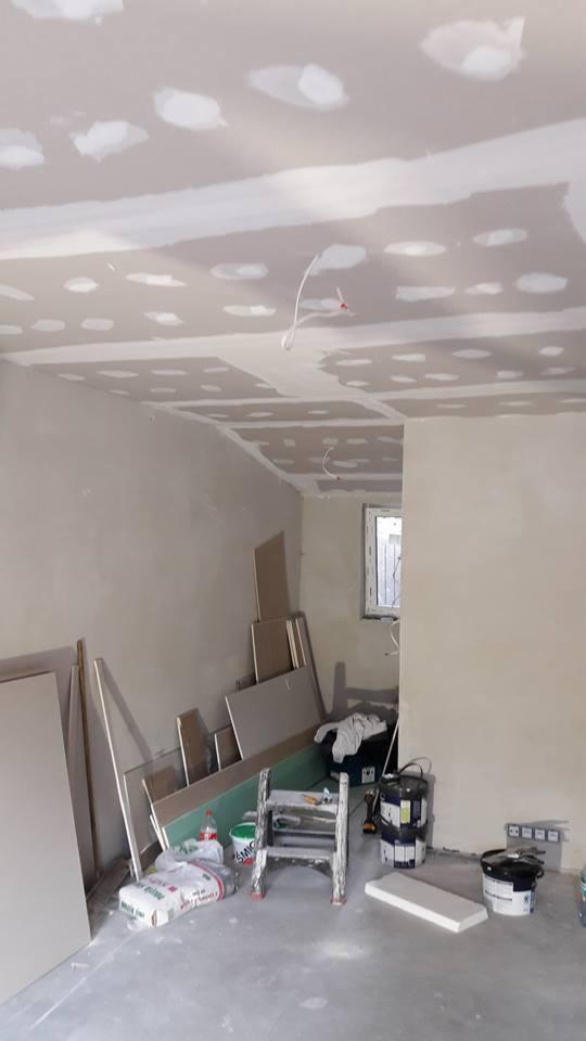 Pokój w trakcie montażu płyt kartonowo-gipsowych na suficie, widoczne łączenia płyt wypełnione masą szpachlową, na podłodze stoją płyty, drabina, worki z zaprawą i puszki z farbą.