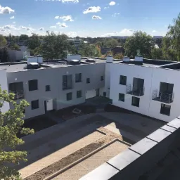 Osiedle KW INWEST: Widok z góry na nowoczesne, dwukondygnacyjne budynki z płaskimi dachami, z elementami obróbki blacharskiej i kominami, otoczone drzewami i brukowanym dziedzińcem.