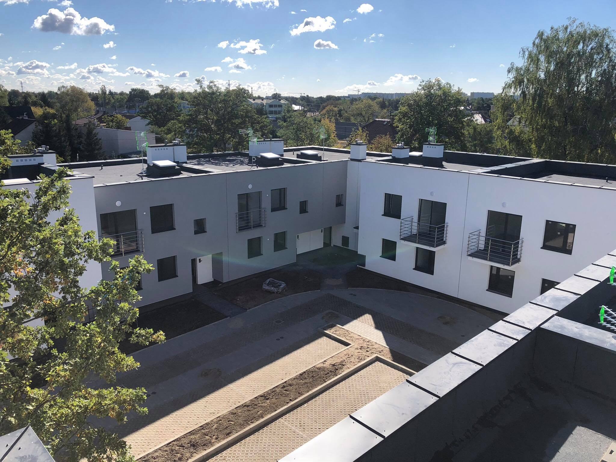 Osiedle KW INWEST: Widok z góry na nowoczesne, dwukondygnacyjne budynki z płaskimi dachami, z elementami obróbki blacharskiej i kominami, otoczone drzewami i brukowanym dziedzińcem.