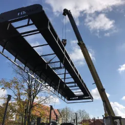 Montaż metalowej konstrukcji z napisem 'TOPACZ' nad zabytkową lokomotywą za pomocą dźwigu BUMAR na tle błękitnego nieba z chmurami.