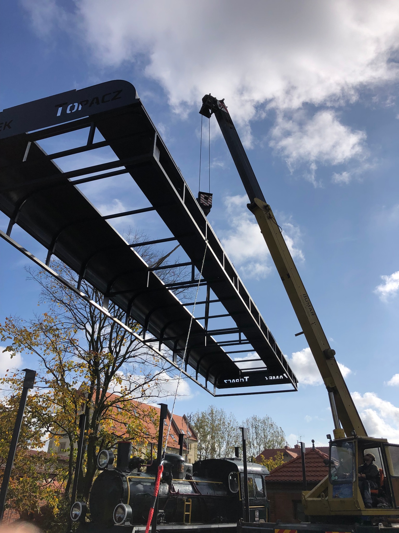 Montaż metalowej konstrukcji z napisem 'TOPACZ' nad zabytkową lokomotywą za pomocą dźwigu BUMAR na tle błękitnego nieba z chmurami.