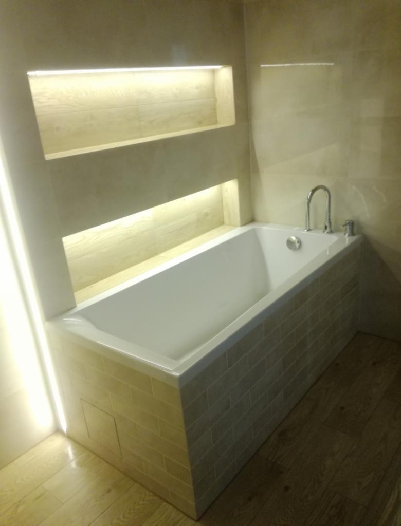 Biała wanna obudowana beżowymi płytkami imitującymi cegłę, z chromowaną armaturą, wbudowane podświetlane półki z jasnymi płytkami, drewnopodobna podłoga, oświetlenie LED wzdłuż ściany.