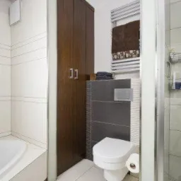 Nowoczesna łazienka z brązową szafą w zabudowie, białą ceramiką i grzejnikiem drabinkowym z ręcznikiem.