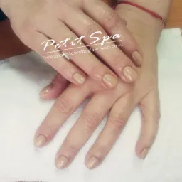 Dłonie po manicure z delikatnym beżowym lakierem, widoczny czerwony sznureczek na nadgarstku, logo 'Petit Spa' nałożone na zdjęcie.