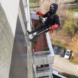 Pracownik w uprzęży, z czerwonymi rękawicami i czarnej odzieży roboczej, zawieszony na linach, wykonuje prace na wysokości na ścianie budynku z balkonami; widok z góry na zaparkowane samochody...