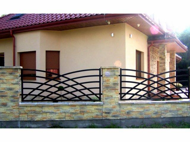 Czarny, metalowy płot o falistym wzorze, osadzony na kamiennym murku przed beżowym domem z brązową dachówką.