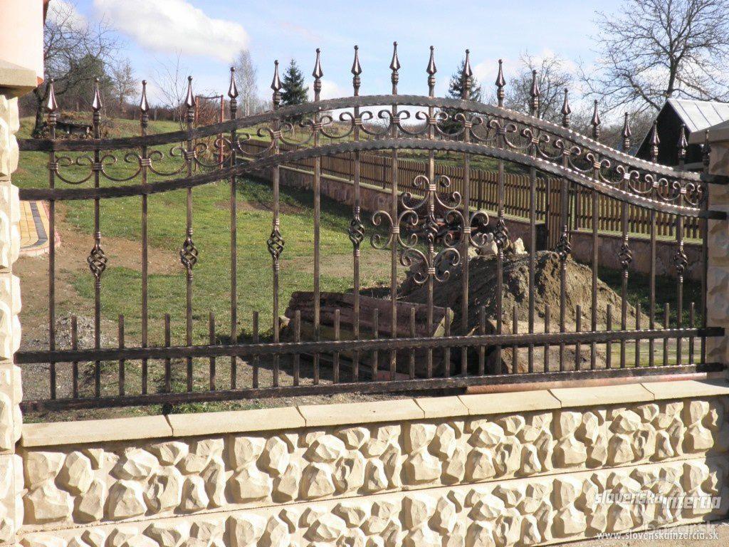 Ozdobne, metalowe przęsło ogrodzenia w kolorze ciemnego brązu z dekoracyjnymi elementami, zamontowane na niskim, kamiennym murku z jasnego piaskowca. W tle widoczny trawnik i częściowo drewniane...