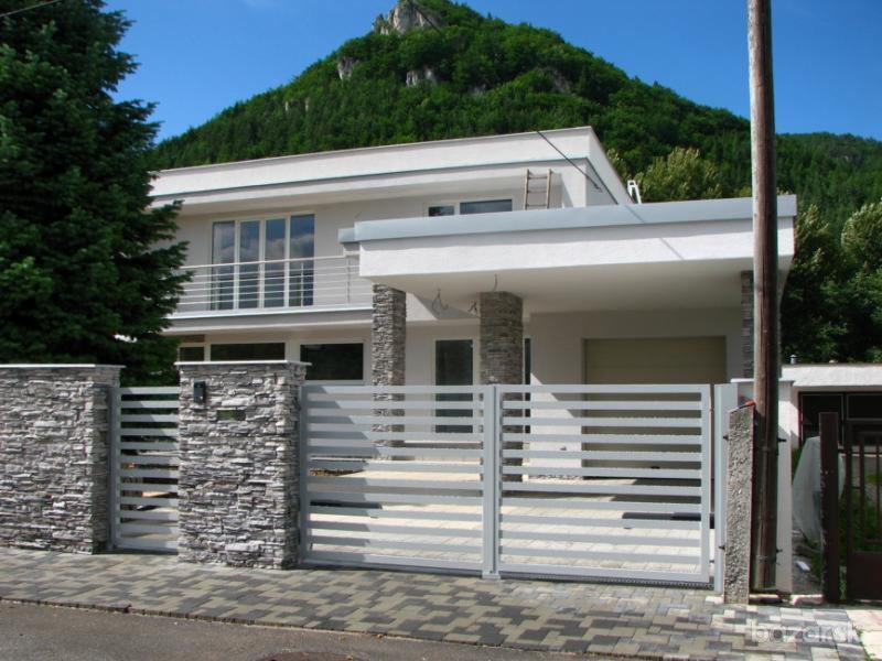 Nowoczesna posesja z szarą, poziomą bramą i ogrodzeniem metalowym z elementami kamiennymi na słupkach, na tle domu i zalesionego wzgórza.