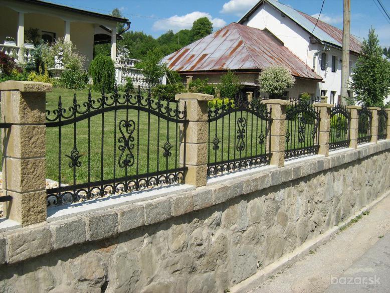 Czarne, ozdobne przęsła z kutymi elementami, osadzone na kamiennych słupkach i murku z nieregularnego kamienia, na tle zielonego trawnika i zabudowań.