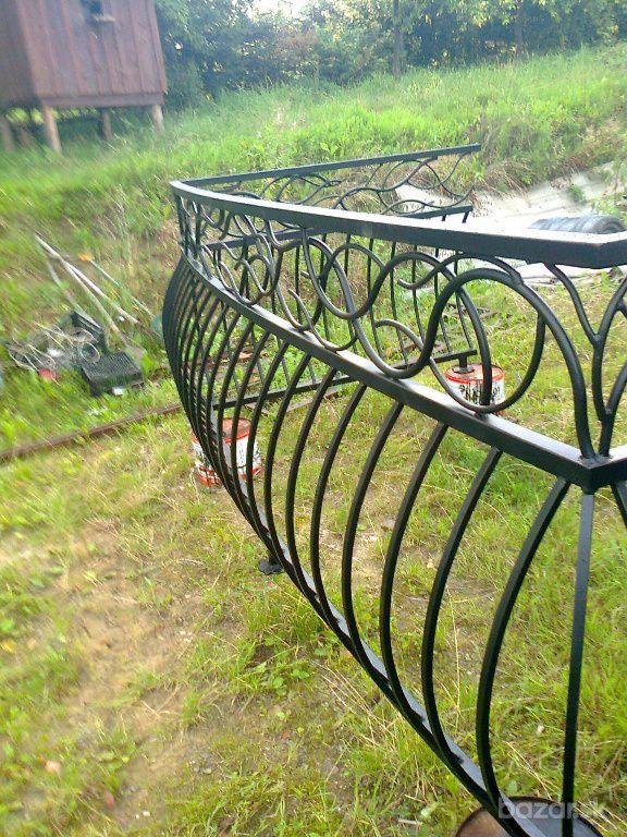 Fragment czarnego, metalowego, ozdobnego balustrady z motywem okręgów, ustawionej na trawniku przed drewnianą budką, z puszką farby widoczną w tle.