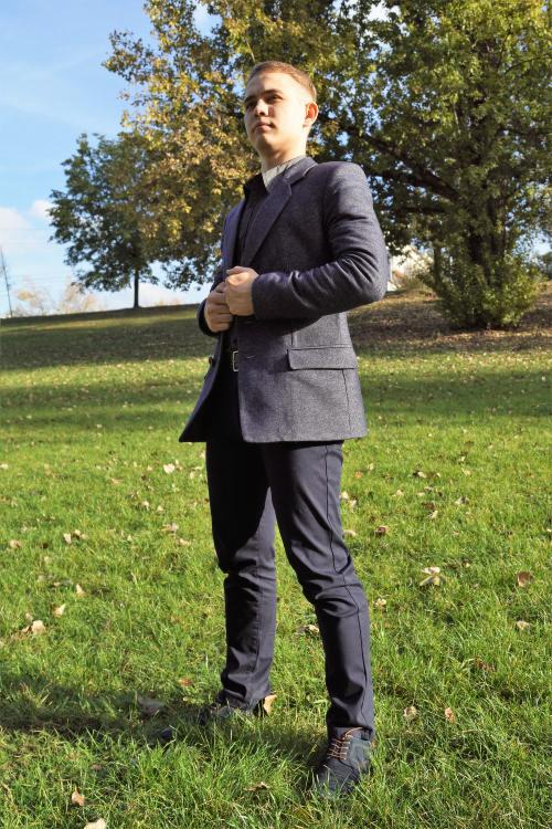 Młody mężczyzna w garniturze z szarego materiału w jodełkę, pozuje na trawie, z zapiętą marynarką i widocznym paskiem, na tle drzew i błękitnego nieba.