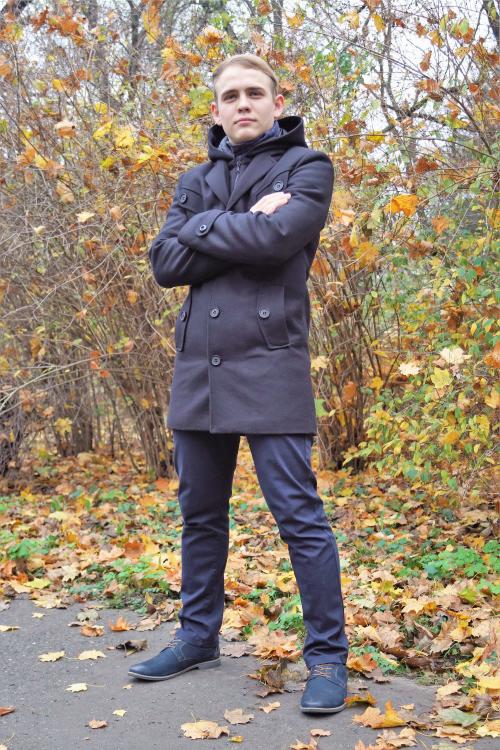 Młody mężczyzna w granatowym płaszczu z kapturem, spodniach i butach, stojący na tle jesiennych krzewów z opadłymi liśćmi.