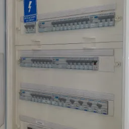 Otwarta, biała skrzynka rozdzielcza z widocznymi bezpiecznikami różnicowoprądowymi, wyłącznikiem głównym, listwą mocy, ogranicznikiem przepięć i licznikiem energii elektrycznej. Widoczne oznaczenia...