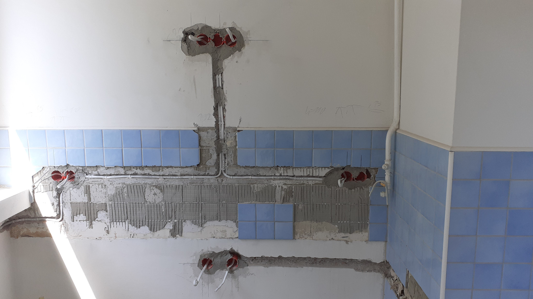 Ściana z częściowo zdjętymi niebieskimi kafelkami, odsłonięte przewody elektryczne i puszki w surowym tynku, widoczne oznaczenia na ścianie ołówkiem.