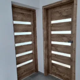 Dwa skrzydła drzwiowe wewnętrzne z drewnopodobną okleiną i poziomymi, prostokątnymi przeszkleniami, umieszczone w białej ścianie, widoczne na szarych płytkach podłogowych, jedno z zamontowaną klamką.