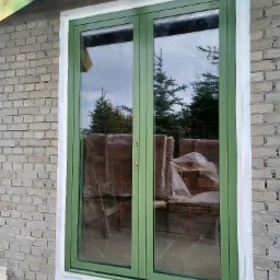 Nowo zamontowane, dwuskrzydłowe okno w zielonej ramie, zabezpieczone folią ochronną na zewnątrz budynku z cegły. W szybie odbijają się drzewa iglaste i pochmurne niebo.