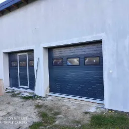 Dwa segmentowe drzwi garażowe w kolorze grafitowym, z małymi oknami, zamontowane w surowej elewacji budynku, widoczne ślady po pracach budowlanych.