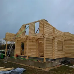 Surowy stan budowy domu z bali, widoczne drewniane ściany, otwory okienne i dach w trakcie wznoszenia, drabina oparta o ścianę, materiały budowlane na trawie, pochmurne niebo w tle.