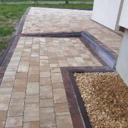 Ułożona nawierzchnia z beżowo-brązowej kostki brukowej z ciemniejszą obwódką, przylegająca do ściany budynku i granicząca z rabatą wypełnioną drobnymi kamieniami.