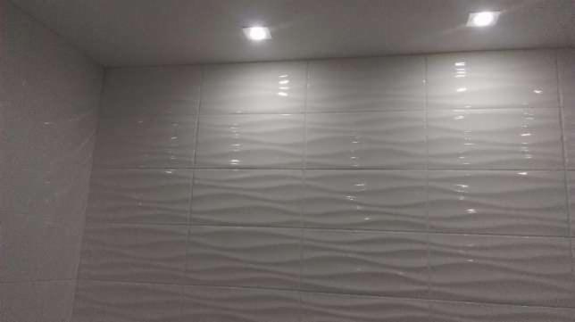 Ściana wyłożona białymi, falistymi płytkami ceramicznymi w łazience, oświetlona dwoma punktowymi lampami sufitowymi.