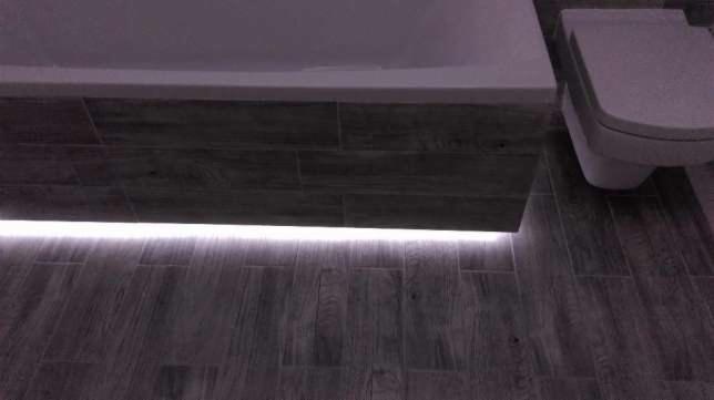 Nowoczesna łazienka z podświetleniem LED pod zabudową wanny i podwieszaną toaletą, płytki imitujące drewno na podłodze i obudowie wanny.