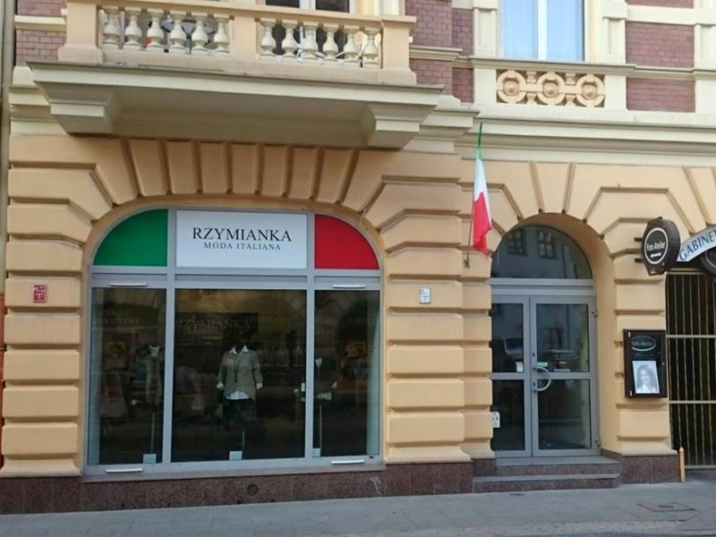 Fasada budynku z butikiem odzieżowym 'Rzymianka Moda Italiana' z logo w kolorach flagi Włoch i włoską flagą powiewającą nad wejściem.