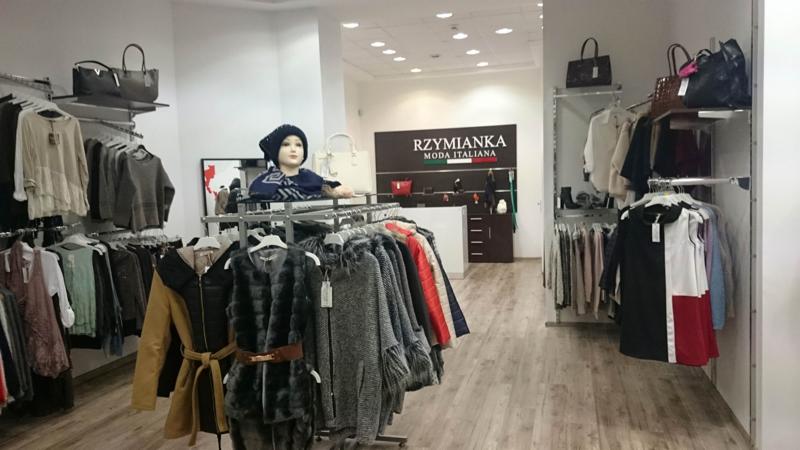 Wnętrze butiku z ubraniami, widoczne wieszaki z różnorodną kolekcją, manekin w niebieskiej czapce i szalu, oraz logo firmy 'RZYMIANKA MODA ITALIANA' w tle.