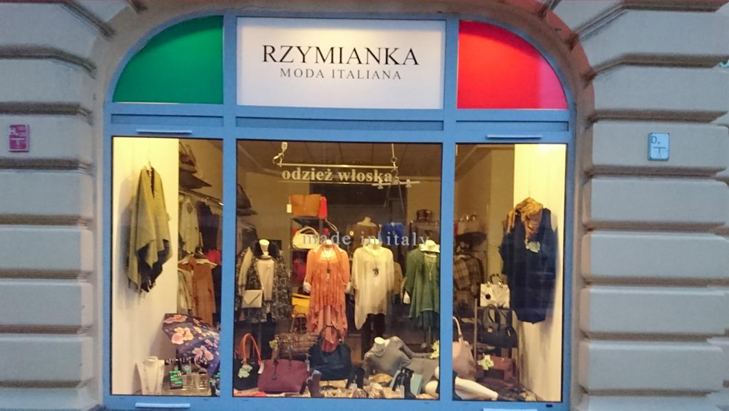 Wystawa butiku z włoską modą, z manekinami prezentującymi kolekcję, torbami i akcesoriami, z szyldem 'Rzymianka Moda Italiana' i flagą włoską w górnej części okna.