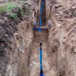 Wykop w ziemi z widocznymi niebieskimi rurami wodociągowymi i złączkami, w tle mała koparka na trawiastym terenie.