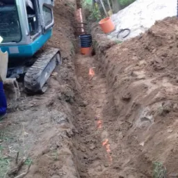 Wykopany rów pod rury kanalizacyjne z ułożonymi odcinkami rur, widoczny minikoparka i hałda ziemi.