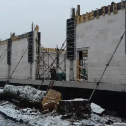 Budowa domu z białych bloczków w zimowej scenerii, widoczne metalowe szalunki okienne i podpory, w tle ścięte drzewa.