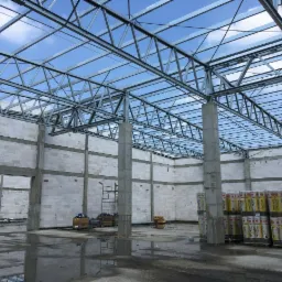 Wnętrze w trakcie budowy: surowe ściany z białego betonu komórkowego, stalowa konstrukcja dachu pod błękitnym niebem, betonowe słupy, materiały izolacyjne w żółtych opakowaniach, mokra posadzka...