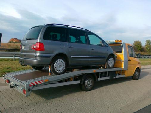 Szary minivan na żółtej lawecie z widocznymi światłami ostrzegawczymi i tablicą rejestracyjną, transportowany po drodze.