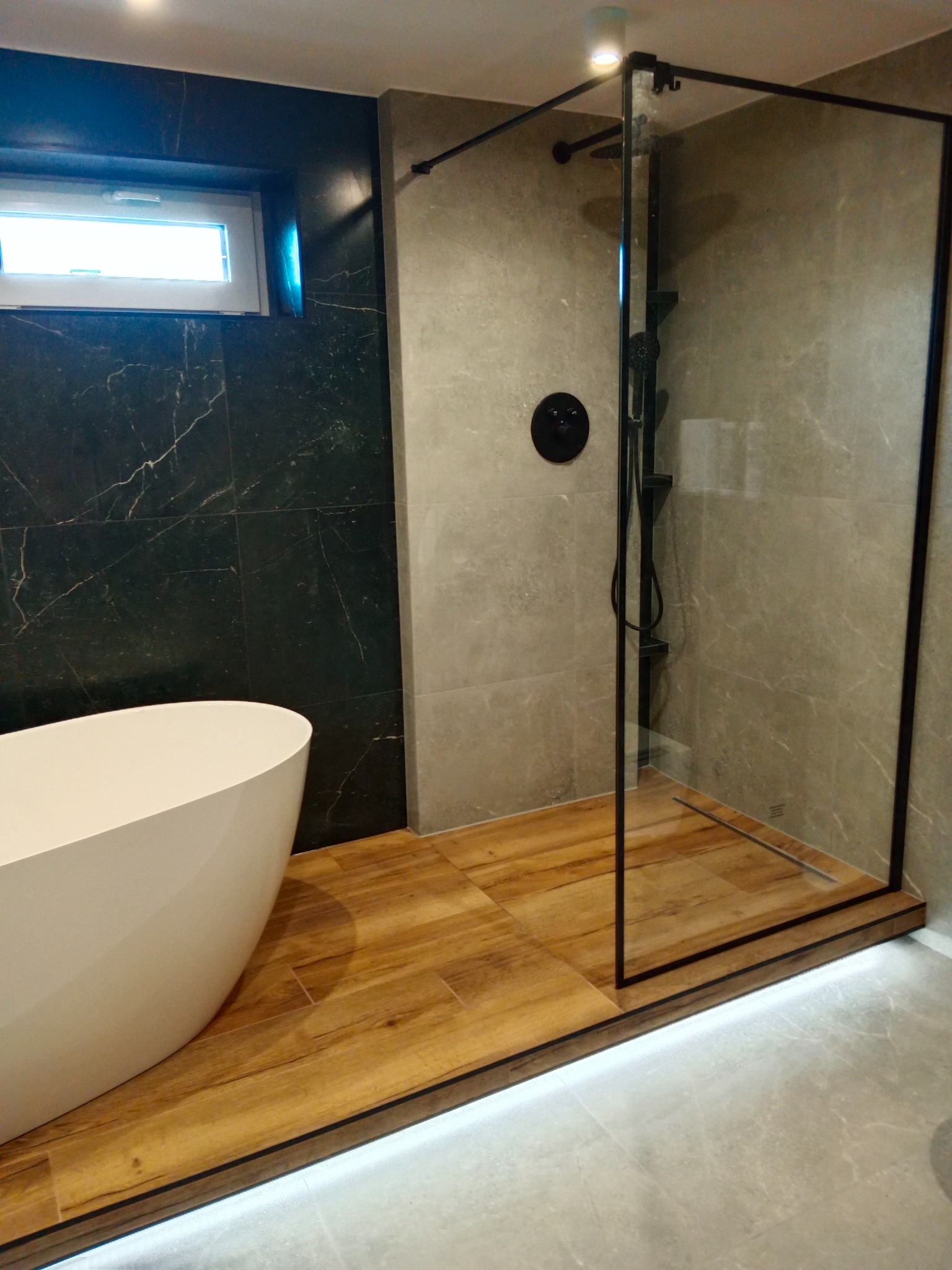 Nowoczesna łazienka z czarnymi akcentami: kabina prysznicowa walk-in z czarną ramą, biała wanna wolnostojąca i marmurowe płytki na ścianach.