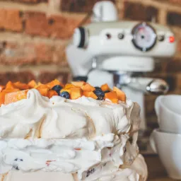 Dwupoziomowy tort bezowy udekorowany kawałkami mango i borówkami, na drewnianym blacie, z mikserem planetarnym w tle i ceglaną ścianą.