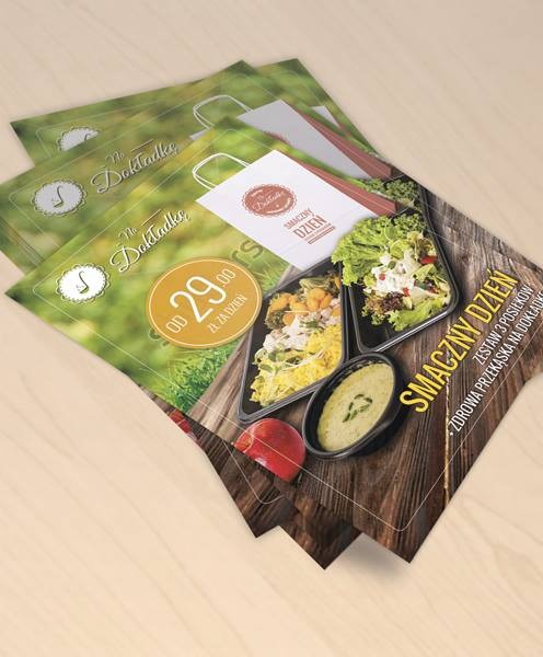 Stos ulotek reklamowych 'Smaczny Dzień' z motywem zdrowego jedzenia w pudełkach i ceną 'od 29 zł za dzień'. Widoczny także papierowa torba z logo.