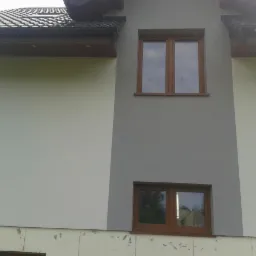 Fragment elewacji budynku z dwoma oknami o drewnianych ramach, gdzie część ściany jest pomalowana na szaro, a część na biało; widoczne niedokończone ocieplenie fundamentów.